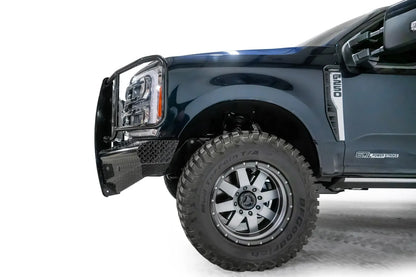 Fab Fours Black Steel front bumper with full guard on a dark blue Ford Super Duty, driver-side profile view of the front corner, showing full grille guard with mesh insert, diamond-plate face panels, the center sensor/plate mount, and angled side wings
