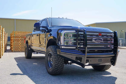 Fab Fours Black Steel front bumper with full guard on a dark blue Ford Super Duty, front passenger-side three-quarter view outdoors, showing full grille guard with mesh insert, diamond-plate face panels, the center sensor/plate mount, and angled side wings