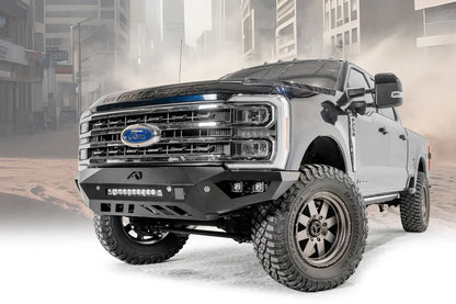 Fab Fours Vengeance front bumper on a gray and dark blue two-tone Ford Super Duty, front driver-side three-quarter view against a dusty city backdrop, showing the sculpted face, integrated LED light bar and cube light pockets