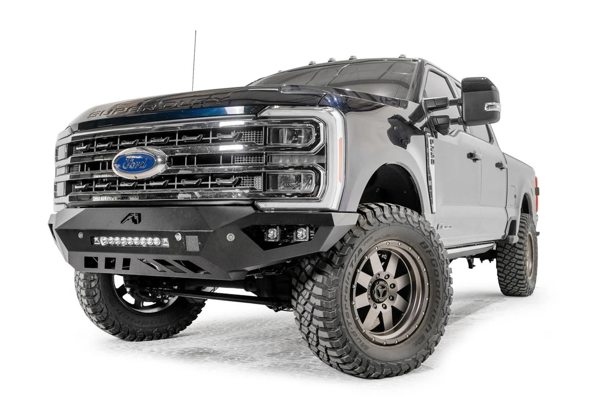 Fab Fours Vengeance front bumper on a gray and dark blue two-tone Ford Super Duty, front driver-side three-quarter view, showing the sculpted no-guard face, integrated LED light bar and cube light pockets