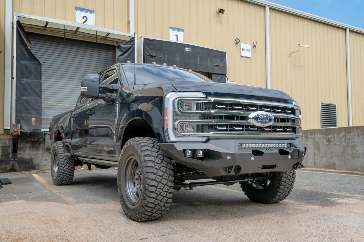 Fab Fours Matrix front bumper on a dark blue Ford Super Duty F-250 outside a warehouse, front passenger-side three-quarter view, showing the no-guard winch bumper, LED light bar and cube lights