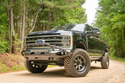 Fab Fours Matrix front bumper with pre-runner guard on a dark blue Ford Super Duty F-250 on a forest trail, front driver-side three-quarter view, showing the pre-runner hoop, LED light bar and cube lights