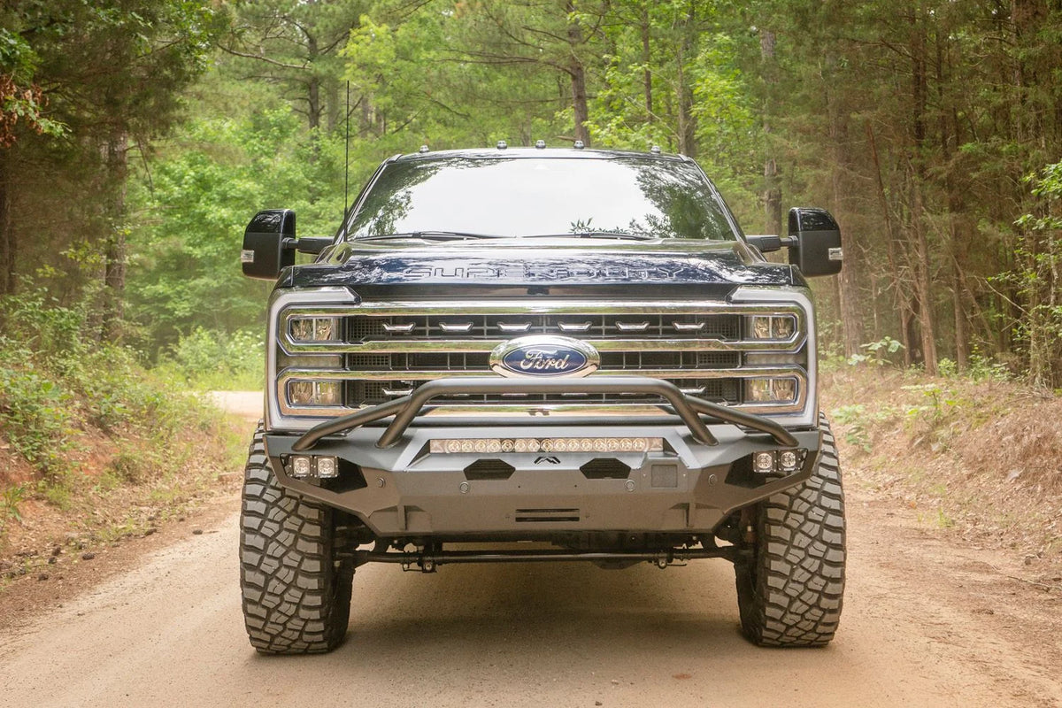 Fab Fours Matrix front bumper with pre-runner guard on a dark blue Ford Super Duty F-250 on a forest trail, straight-on front view, showing the pre-runner hoop, LED light bar and winch opening