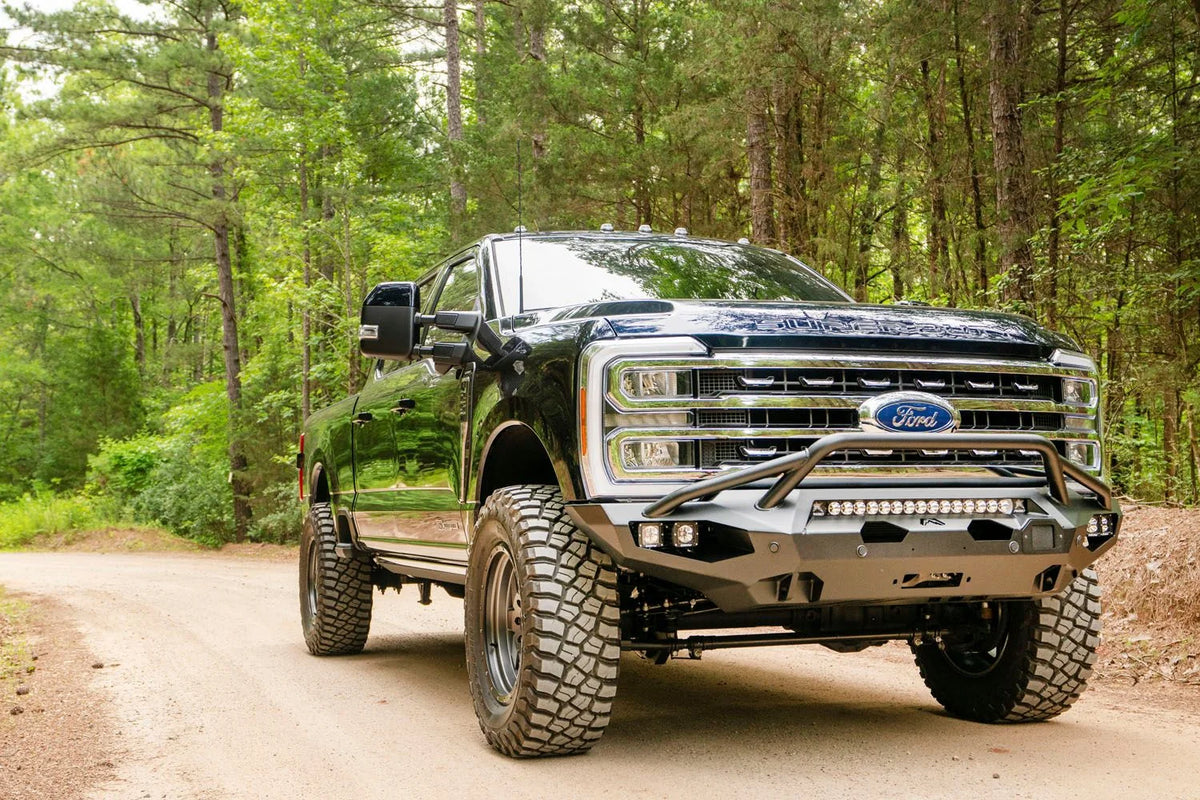 Fab Fours Matrix front bumper with pre-runner guard on a dark blue Ford Super Duty F-250 on a dirt road, front passenger-side three-quarter view, showing the pre-runner hoop, LED light bar and cube lights