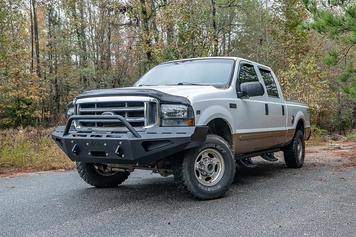 Fab Fours Red Steel front bumper with pre-runner guard on a white and tan two-tone Ford Super Duty F-250 on a wooded road, front driver-side three-quarter view, showing the pre-runner hoop, angular bumper face and wing profile