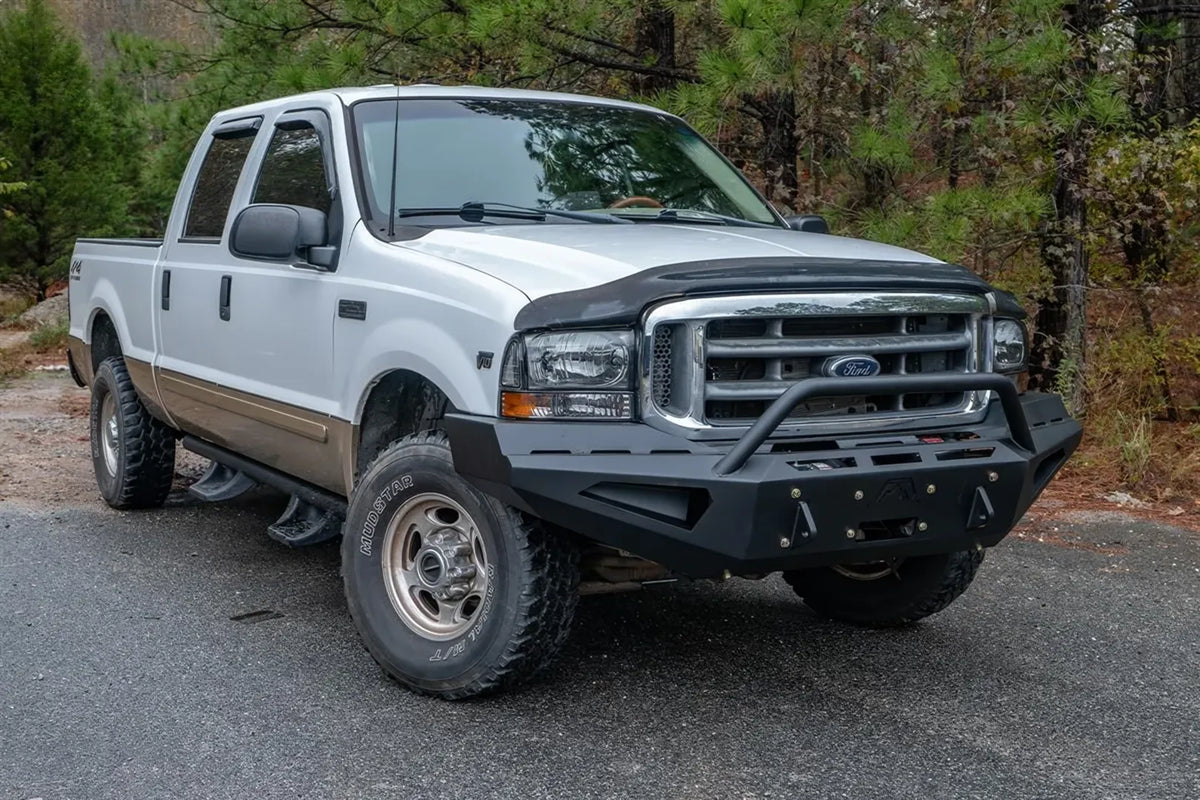 Fab Fours Red Steel front bumper with pre-runner guard on a white and tan two-tone Ford Super Duty F-250 parked on a wooded road, front passenger-side three-quarter view, showing the pre-runner hoop, angular bumper face and side light cutouts