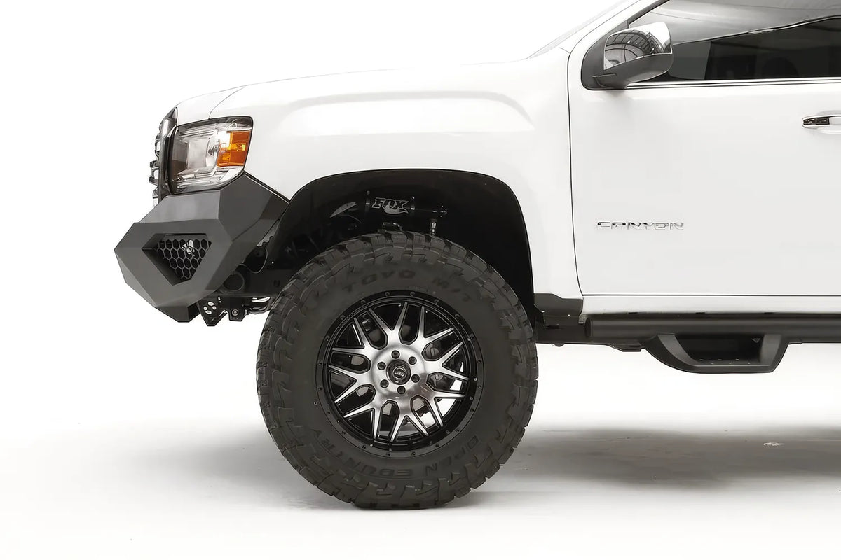 Fab Fours Vengeance front bumper on a white GMC Canyon, close-up of the driver-side corner, showing the bumper wing, honeycomb light pocket and wheel-well clearance