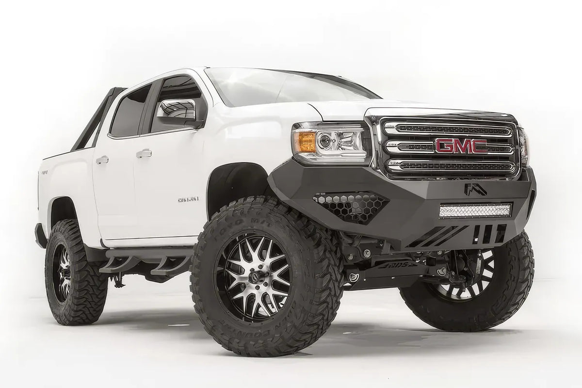 Fab Fours Vengeance front bumper on a white GMC Canyon, front driver-side three-quarter view, showing the sculpted no-guard face, LED light bar and angular skid section
