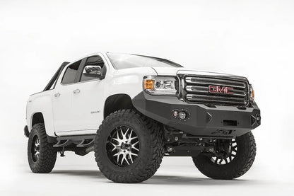 Fab Fours Premium front bumper on a white GMC Canyon, front driver-side three-quarter view, showing the no-guard winch bumper face, round fog light cutouts and wrapped wings