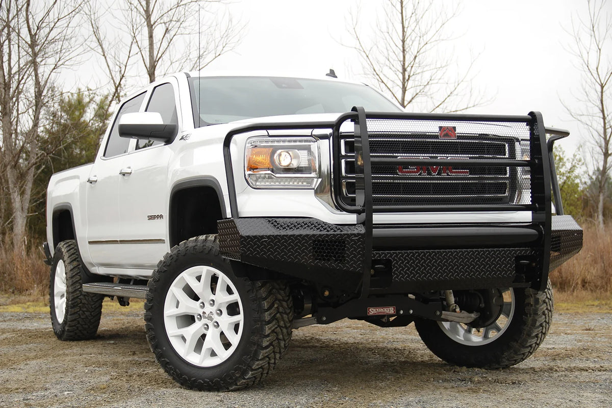 Fab Fours Black Steel front bumper with full guard on a white GMC Sierra 1500 parked on gravel, front passenger-side three-quarter view, showing the full grille guard, headlight hoops and diamond-plate bumper face