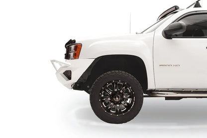 Fab Fours Vengeance front bumper with pre-runner guard on a white GMC Sierra 2500HD, driver-side corner profile view, showing the pre-runner tube end, bumper wing and honeycomb light pocket
