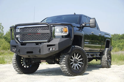 Fab Fours Premium Heavy Duty winch front bumper with full guard on a black GMC Sierra 2500HD parked on gravel, front driver-side three-quarter view, showing the tubular grille guard hoop, winch opening and round fog light cutouts