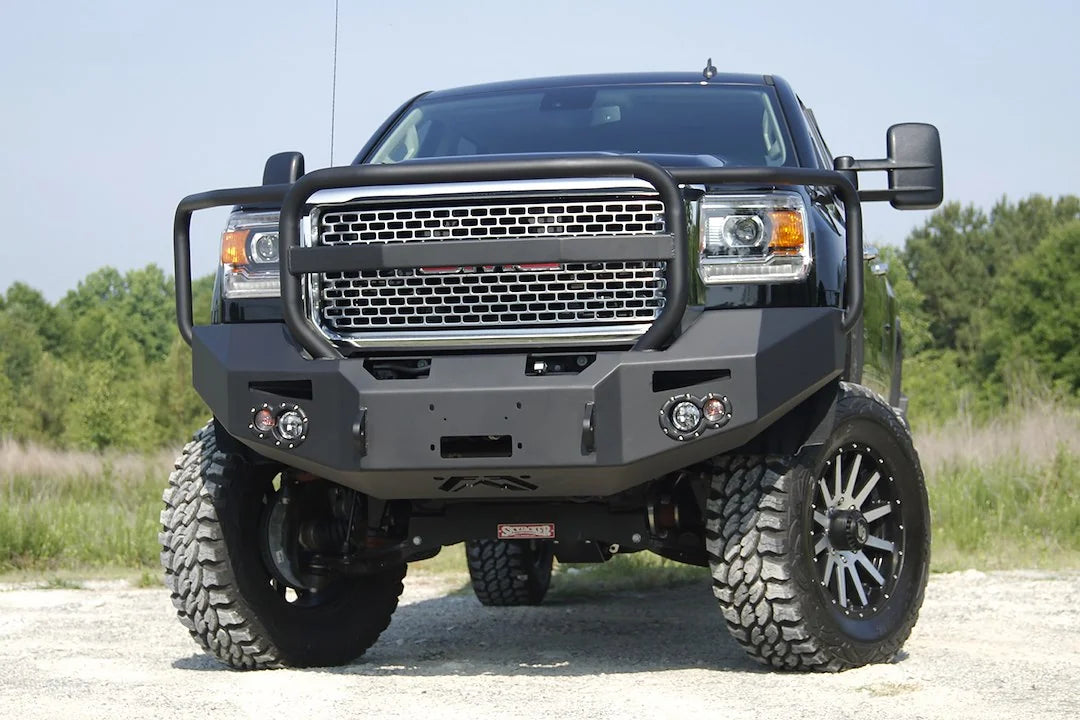 Fab Fours Premium Heavy Duty winch front bumper with full guard on a black GMC Sierra 2500HD parked on gravel, straight-on front view, showing the tubular grille guard hoop, center winch opening and round fog light cutouts