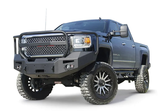 Fab Fours Premium Heavy Duty winch front bumper with full guard on a black GMC Sierra 2500HD, low outdoor close-up of the passenger-side corner, showing the guard hoop, winch fairlead opening and round fog light cutout