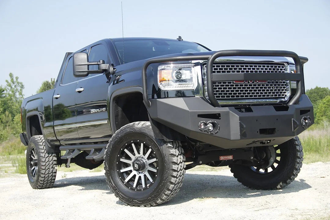 Fab Fours Premium Heavy Duty winch front bumper with full guard on a black GMC Sierra 2500HD parked on gravel, front passenger-side three-quarter view, showing the grille guard hoop, winch opening and fog light cutouts