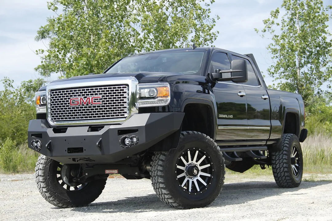 Fab Fours Premium Heavy Duty winch front bumper on a black GMC Sierra 2500HD parked on gravel, front driver-side three-quarter view, showing the no-guard winch bumper face, round fog light cutouts and shackle tabs