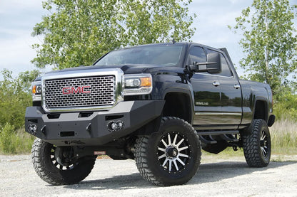 Fab Fours Premium Heavy Duty winch front bumper on a black GMC Sierra 2500HD parked on gravel, front driver-side three-quarter view, showing the no-guard winch bumper face, round fog light cutouts and shackle tabs