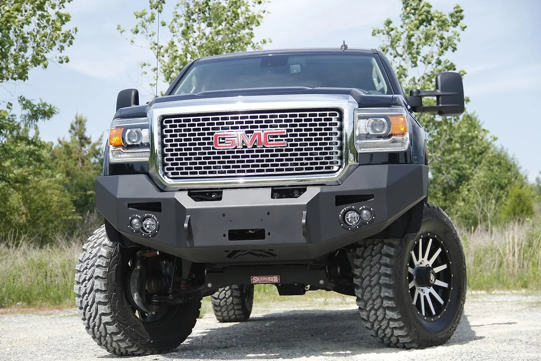 Fab Fours Premium Heavy Duty winch front bumper on a black GMC Sierra 2500HD parked on gravel, straight-on front view, showing the no-guard winch bumper face, center winch opening and round fog light cutouts