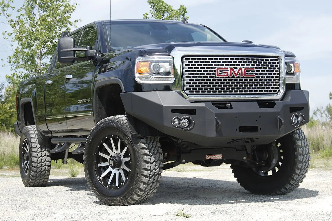 Fab Fours Premium Heavy Duty winch front bumper on a black GMC Sierra 2500HD parked on gravel, front passenger-side three-quarter view, showing the no-guard winch bumper face, round fog light cutouts and wrapped wings