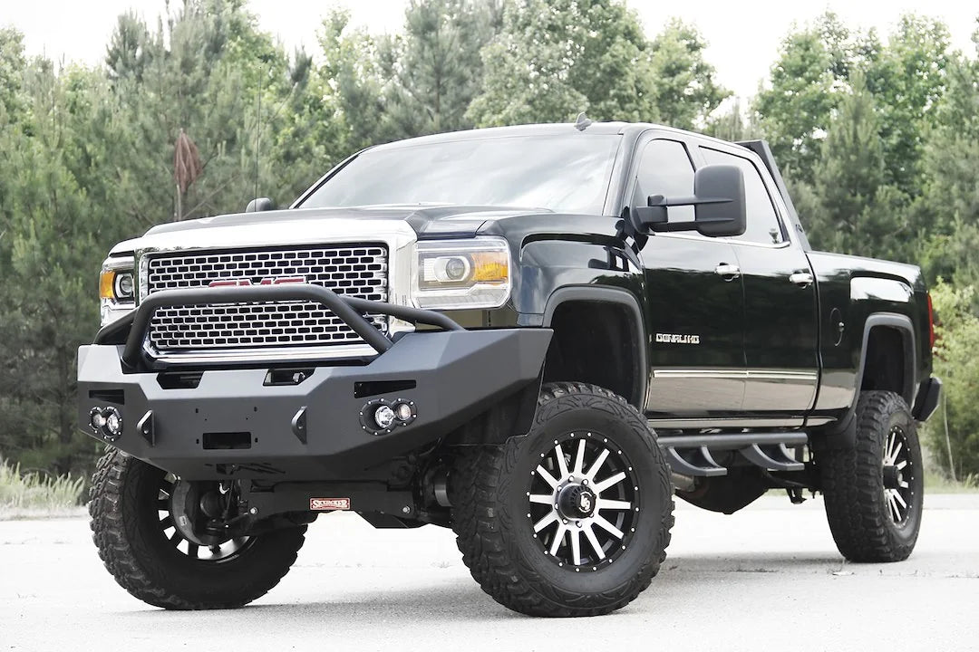 Fab Fours Premium Heavy Duty winch front bumper with pre-runner guard on a black GMC Sierra 2500HD on a paved lot, front driver-side three-quarter view, showing the pre-runner hoop, winch opening and round fog light cutouts