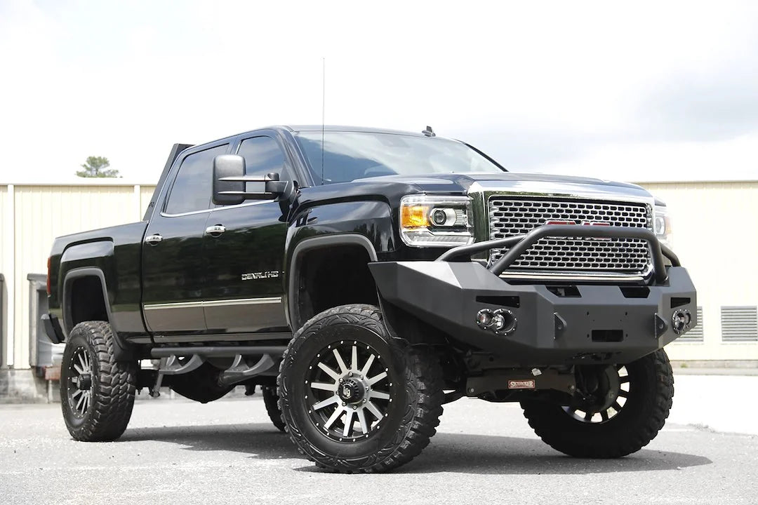 Fab Fours Premium Heavy Duty winch front bumper with pre-runner guard on a black GMC Sierra 2500HD parked outside a building, front passenger-side three-quarter view, showing the pre-runner hoop, winch opening and round fog light cutouts