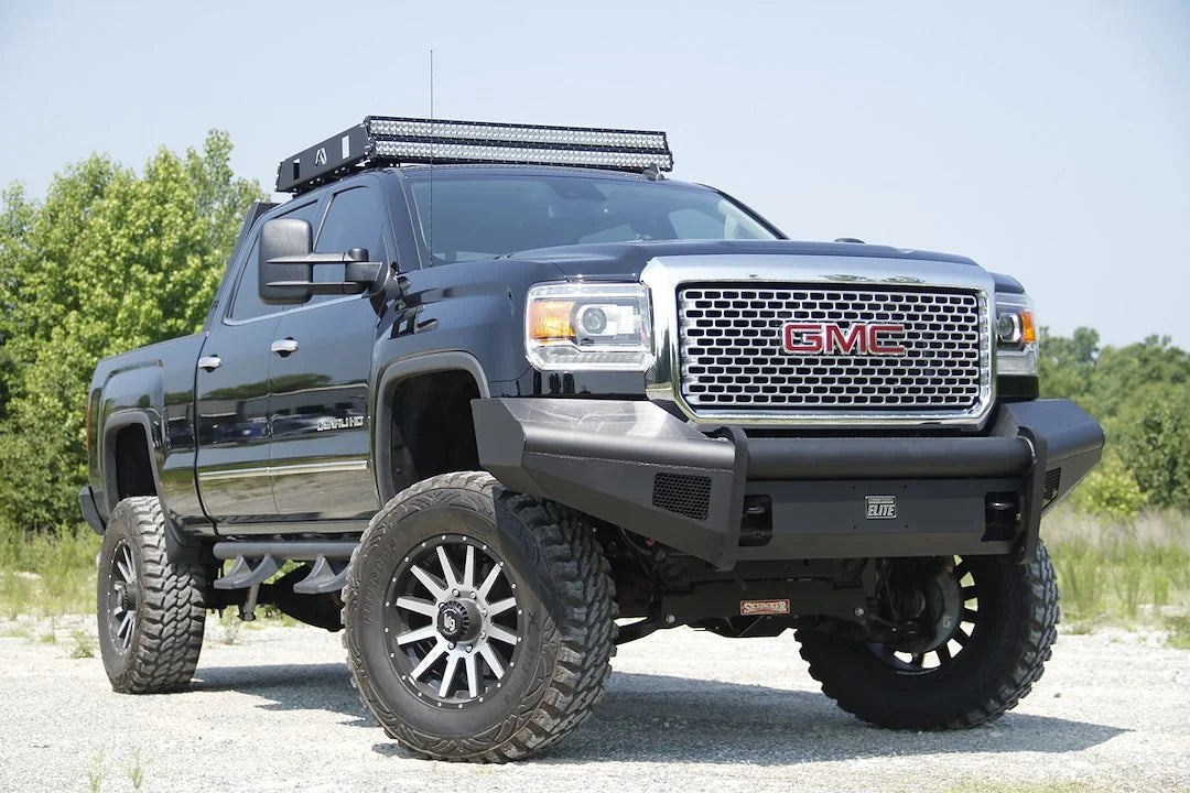 Fab Fours Black Steel Elite front bumper on a black GMC Sierra 2500HD parked on gravel, front passenger-side three-quarter view, showing the no-guard smooth-plate face, Elite badge and wrapped wings