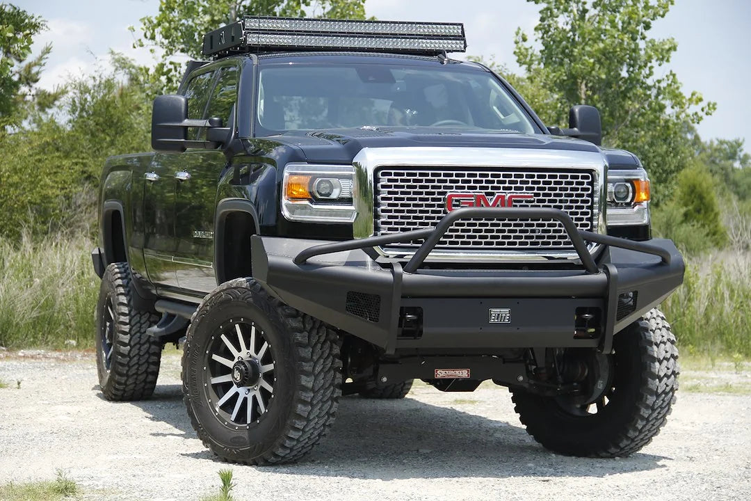 Fab Fours Black Steel Elite front bumper with pre-runner guard on a black GMC Sierra 2500HD parked on gravel, front passenger-side three-quarter view, showing the pre-runner hoop, smooth-plate face and Elite badge