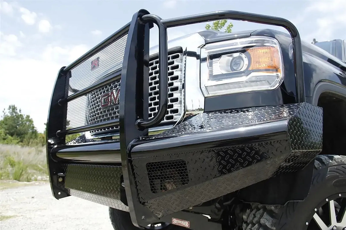 Fab Fours Black Steel front bumper with full guard on a black GMC Sierra 2500HD, low outdoor close-up of the driver-side corner, showing the guard uprights, mesh grille insert and diamond-plate wing
