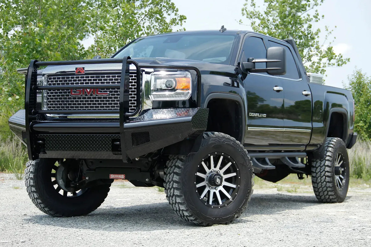 Fab Fours Black Steel front bumper with full guard on a black GMC Sierra 2500HD parked on gravel, front driver-side three-quarter view, showing the full grille guard, mesh grille insert and diamond-plate bumper face