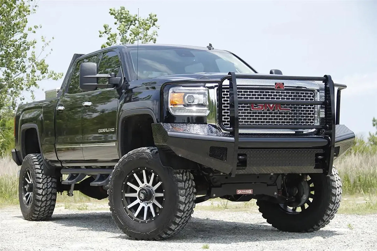 Fab Fours Black Steel front bumper with full guard on a black GMC Sierra 2500HD parked on gravel, front passenger-side three-quarter view, showing the full grille guard, headlight hoops and diamond-plate wings