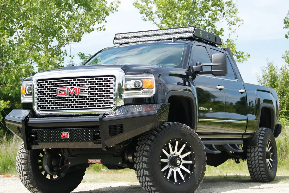 Fab Fours Black Steel front bumper on a black GMC Sierra 2500HD parked on gravel, front driver-side three-quarter view, showing the no-guard diamond-plate face, top tube and wrapped wings