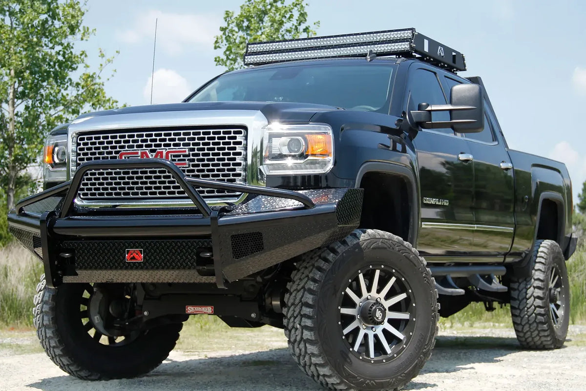 Fab Fours Black Steel front bumper with pre-runner guard on a black GMC Sierra 2500HD parked on gravel, front driver-side three-quarter view, showing the pre-runner hoop, diamond-plate face and wrapped wings