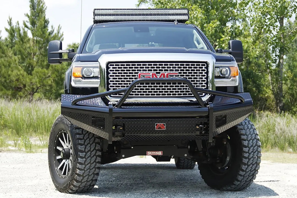 Fab Fours Black Steel front bumper with pre-runner guard on a black GMC Sierra 2500HD parked on gravel, straight-on front view, showing the pre-runner hoop, diamond-plate face and center logo plate