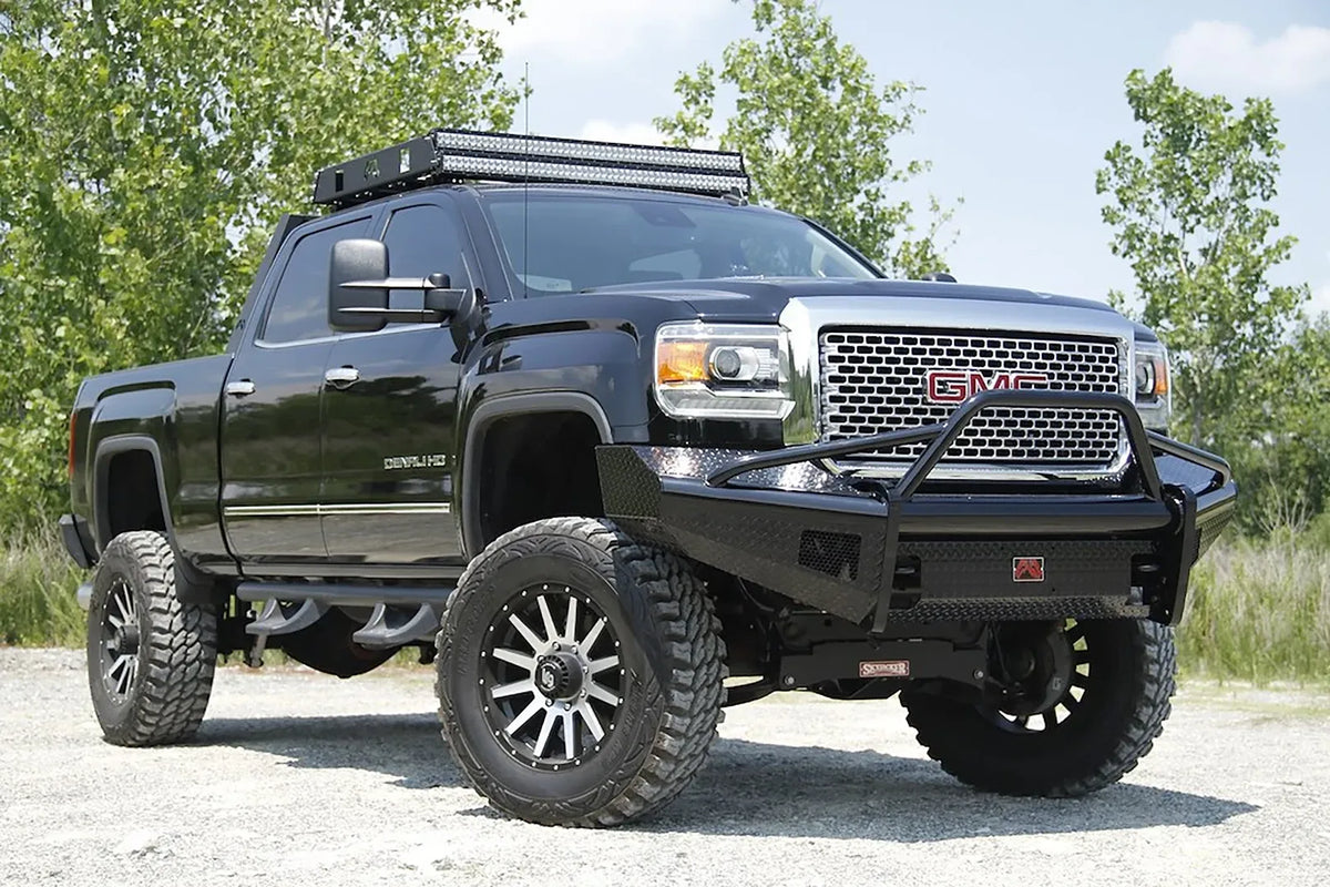 Fab Fours Black Steel front bumper with pre-runner guard on a black GMC Sierra 2500HD parked on gravel, front passenger-side three-quarter view, showing the pre-runner hoop, diamond-plate face and wrapped wings