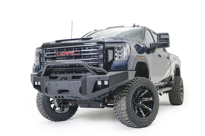 Fab Fours Premium Winch front bumper with pre-runner guard on a blue and gray GMC Sierra 2500HD/3500HD, front driver-side three-quarter view, showing pre-runner hoop, winch mount opening, cube light pods