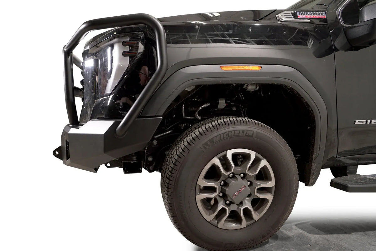 Fab Fours Premium Winch front bumper with full guard on a black GMC Sierra 2500HD/3500HD, close-up of the front corner, showing full guard side hoop, bumper end cap, wheel-well clearance