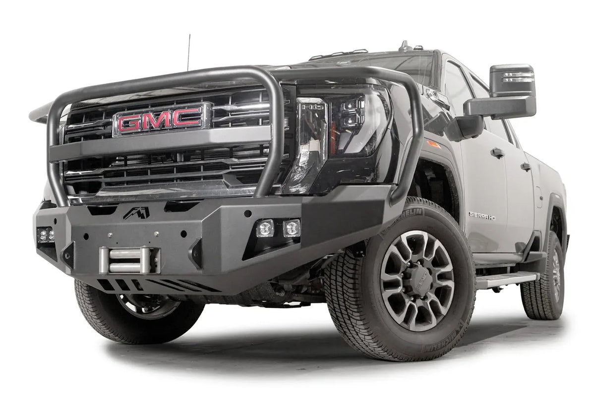 Fab Fours Premium Winch front bumper with full guard on a black GMC Sierra 2500HD/3500HD, front driver-side three-quarter view, showing full guard hoop, winch mount, cube light pods, angled end caps