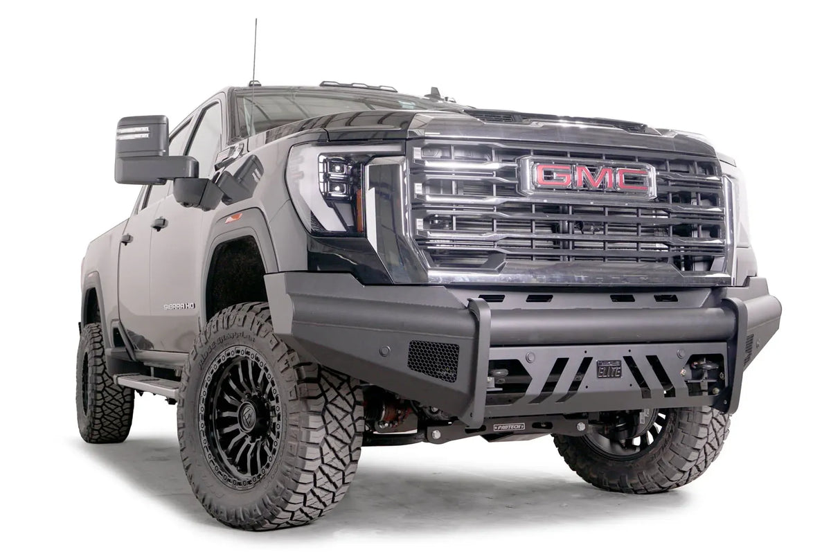 Fab Fours Black Steel Elite front bumper on a black GMC Sierra 2500HD/3500HD, front passenger-side three-quarter view, showing Elite badge, slotted skid plate, mesh end inserts, top tube
