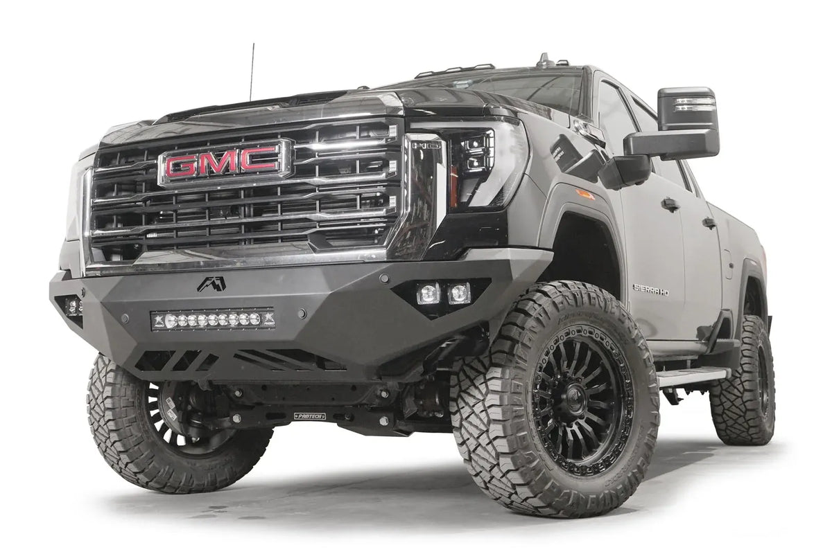 Fab Fours Vengeance front bumper on a black GMC Sierra 2500HD/3500HD, front driver-side three-quarter view, showing integrated light bar, cube light pods, angular skid plate