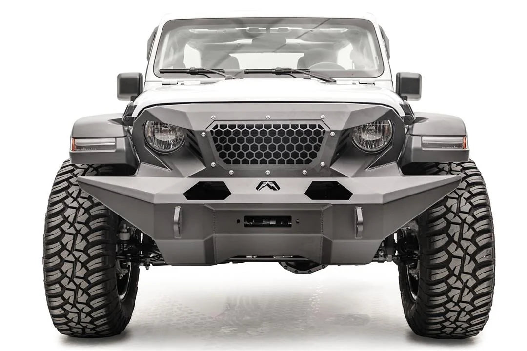 Fab Fours full-width Grumper front bumper on a lifted white Jeep Wrangler JL Rubicon, straight-on front view, showing the wide faceted bumper wings that extend past the tires, the honeycomb mesh grille guard, Fab Fours logo and D-ring mounts
