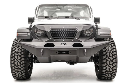 Fab Fours full-width Grumper front bumper on a lifted white Jeep Wrangler JL Rubicon, straight-on front view, showing the wide faceted bumper wings that extend past the tires, the honeycomb mesh grille guard, Fab Fours logo and D-ring mounts