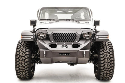 Fab Fours Grumper front winch bumper on a lifted white Jeep Wrangler JL Rubicon, straight-on front view, showing the compact bumper with winch fairlead opening, honeycomb mesh grille guard, Fab Fours logo and D-ring mounts