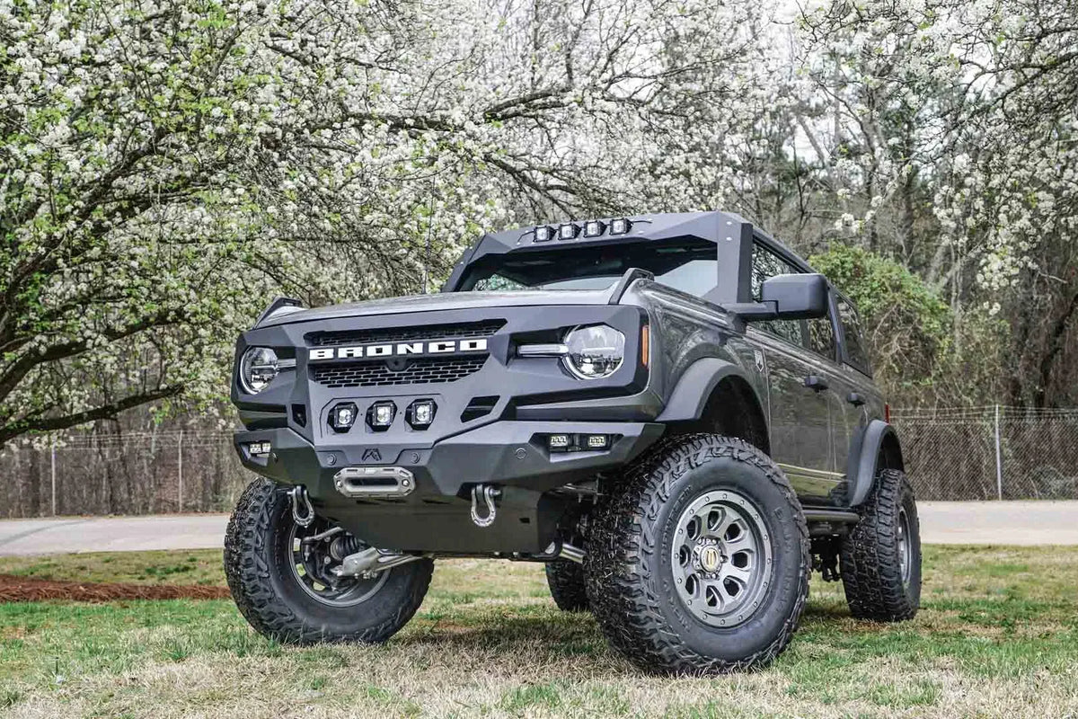 Fab Fours Grumper front bumper on a lifted gray Ford Bronco under flowering trees, driver-side front three-quarter view, with cube lights and D-ring shackles