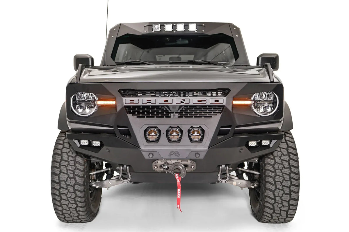Fab Fours Grumper front bumper on a lifted gray Ford Bronco, front view, with three center pod lights, cube lights, Warn winch, and D-ring shackles