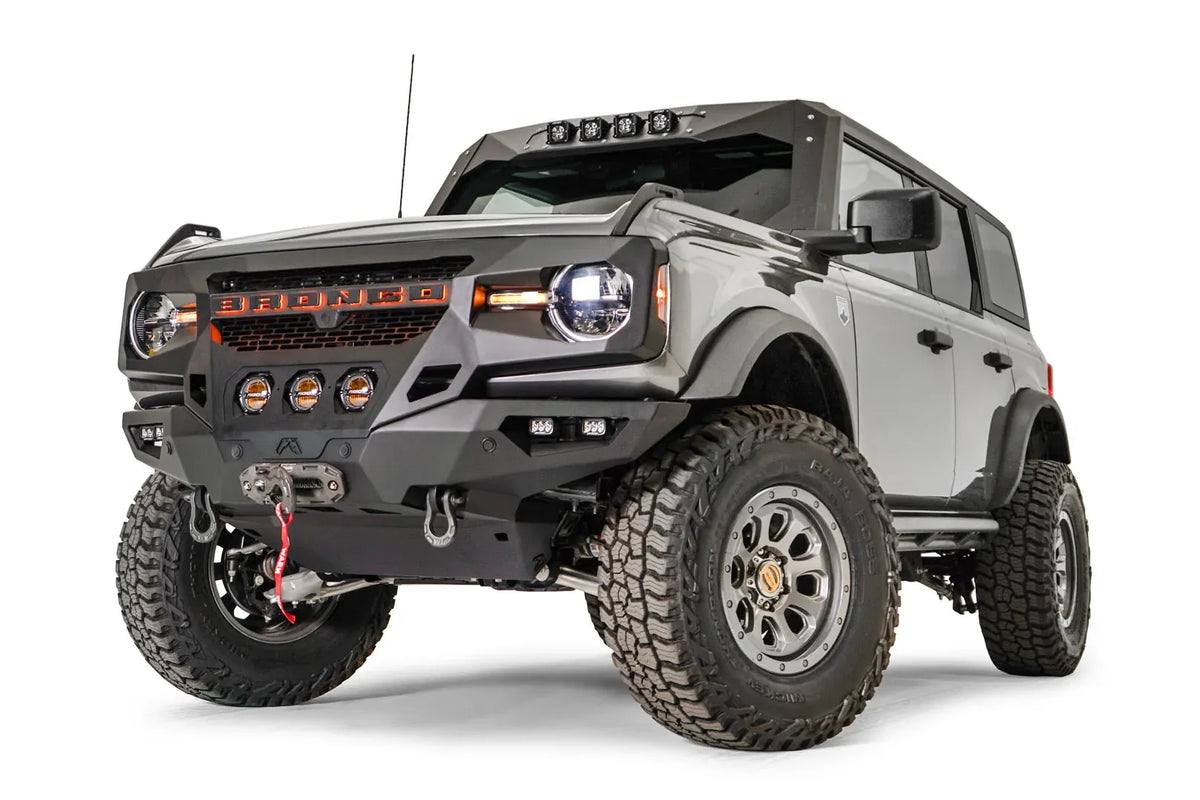 Fab Fours Grumper front bumper with three center pod lights on a lifted gray Ford Bronco, driver-side front three-quarter view, with Warn winch and D-ring shackles