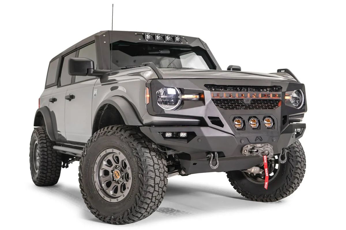 Fab Fours Grumper front bumper with three center pod lights on a lifted gray Ford Bronco, passenger-side front three-quarter view, with Warn winch and D-ring shackles