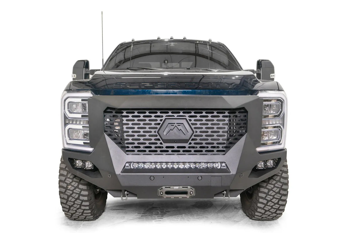 Fab Fours Grumper front winch bumper on a dark blue 2023+ Ford Super Duty, straight-on front view, showing the full-width slotted grille with the hex Fab Fours badge, the center LED light bar, pod lights on each side and the winch fairlead