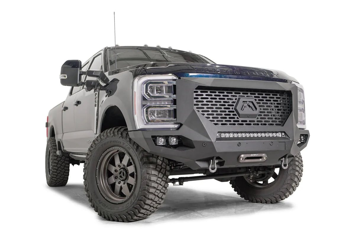 Fab Fours Grumper front winch bumper on a lifted dark blue 2023+ Ford Super Duty, front passenger-side view, showing the slotted grille panel, the lower LED light bar, cube light pockets and the winch fairlead between the D-ring shackles