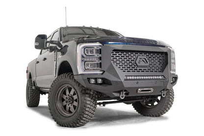 Fab Fours Grumper front winch bumper on a lifted dark blue 2023+ Ford Super Duty, front passenger-side view, showing the slotted grille panel, the lower LED light bar, cube light pockets and the winch fairlead between the D-ring shackles