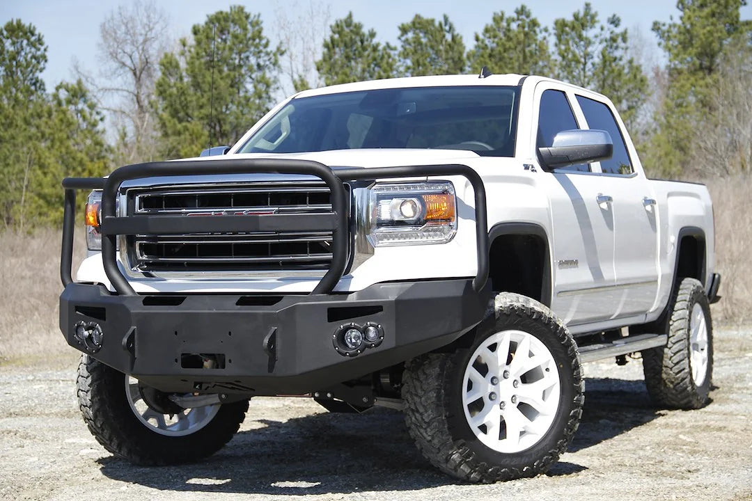 Fab Fours Premium Winch front bumper with full guard on a white GMC Sierra 1500, front driver-side three-quarter view outdoors, showing full guard hoop, winch plate, round fog light pods
